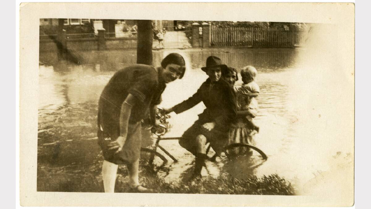 1920s photo of a boy on a bicycle and three other children playing on the edge of Albury floodwaters in the foreground. Another boy is standing by a gate on the other side of the street in the background. Picture: ALBURYCITY COLLECTION